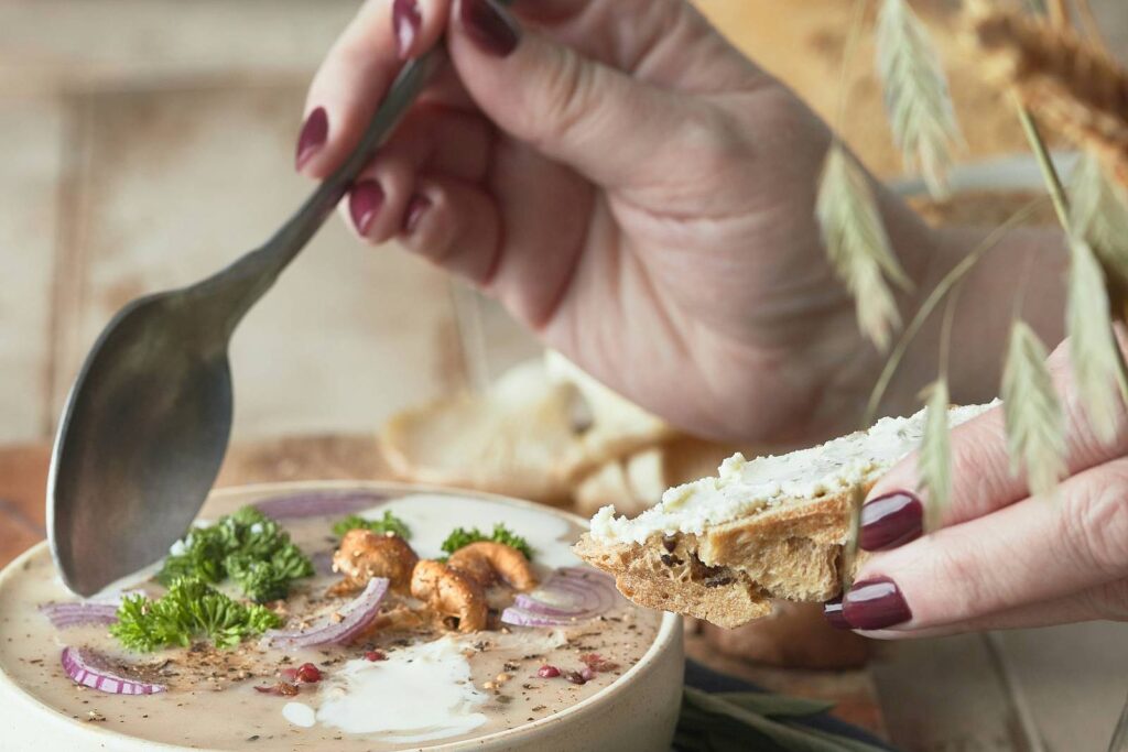 A hand lifting a spoon over a bowl of soup with bread and wheat stalks.