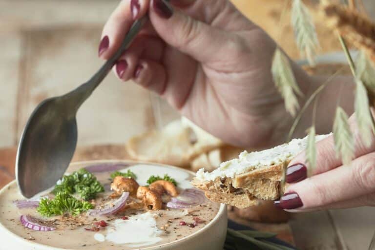 A hand lifting a spoon over a bowl of soup with bread and wheat stalks.