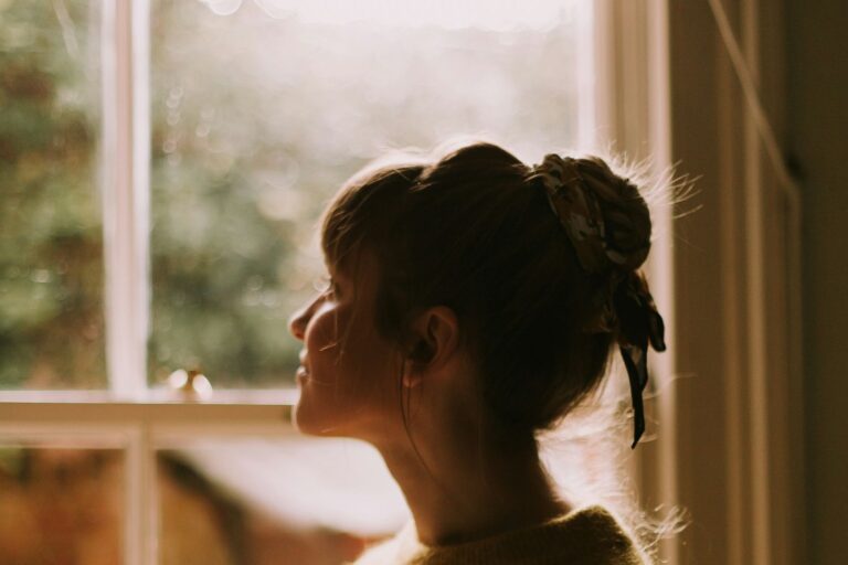 A woman holding a warm mug, looking out of a sunlit window.