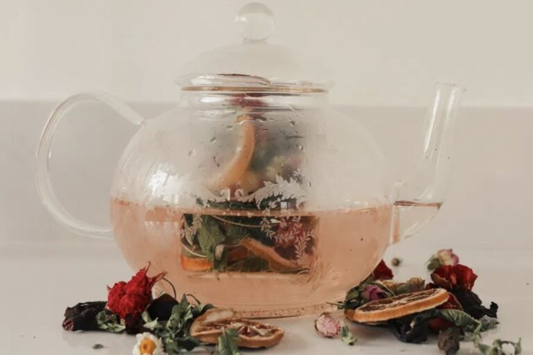 A glass teapot of rose-gold herbal tea surrounded by dried flowers and citrus.