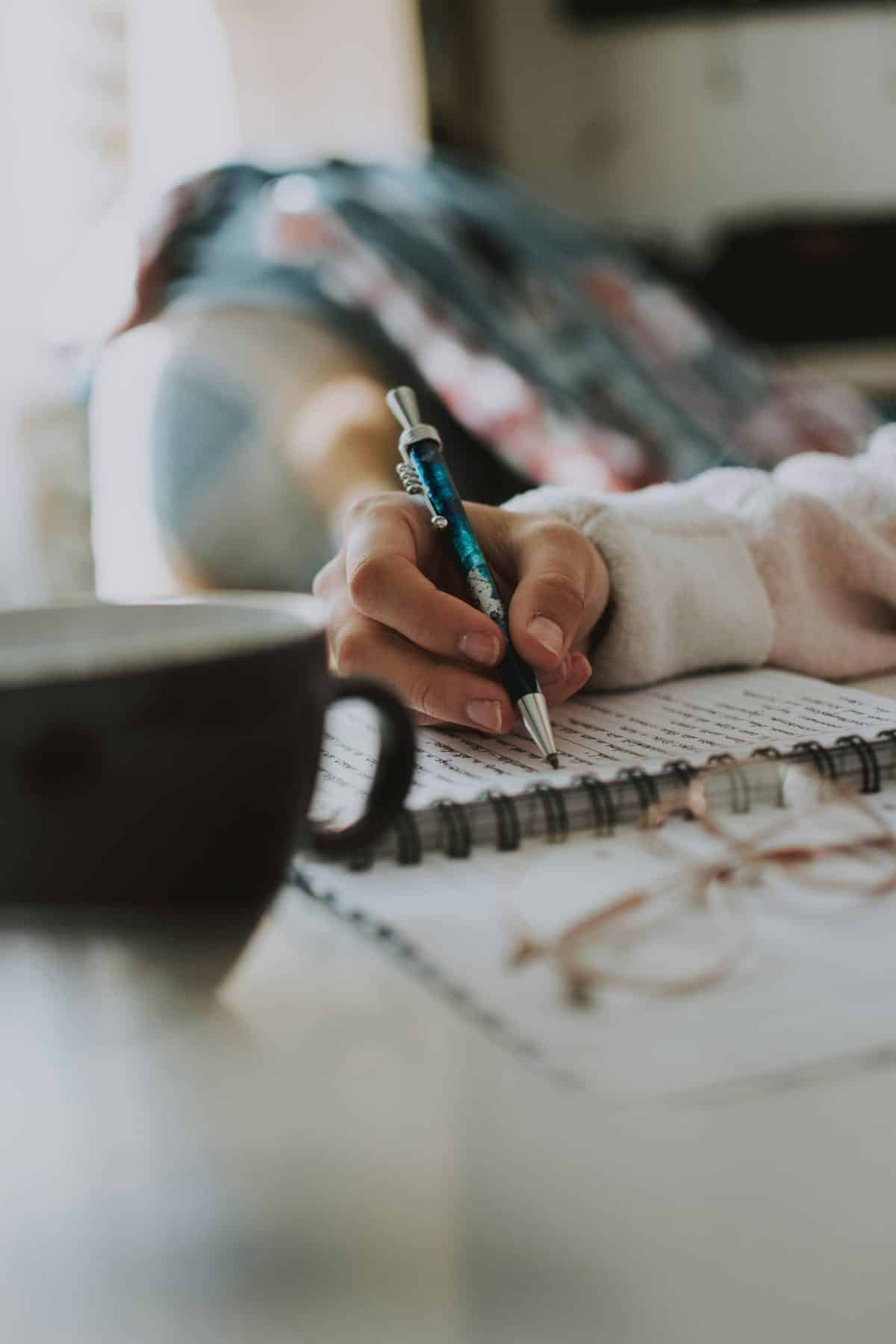 A woman writing quietly in a notebook beside a cup of coffee.