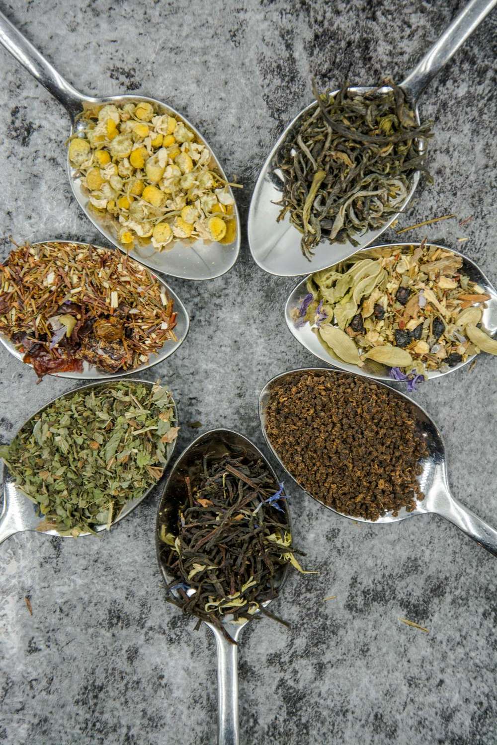 Six spoonfuls of different loose-leaf herbal teas arranged in a circle.