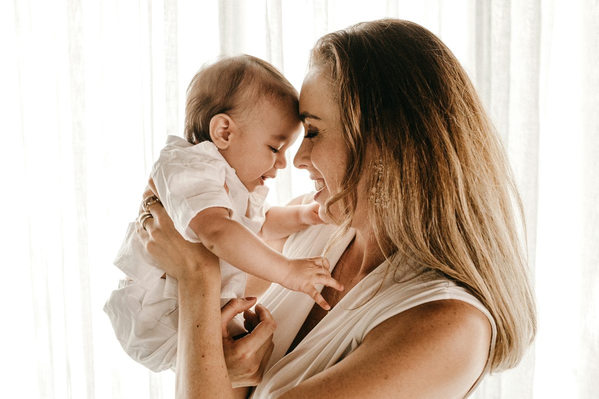 A mother lifting her baby up towards her face in bright window light.