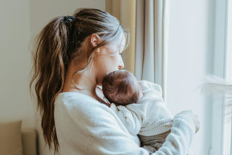 A mother holding her newborn baby by a window in soft natural light.
