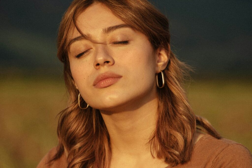A woman standing outdoors in soft daylight with her eyes closed.