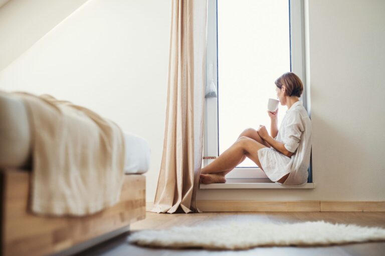 A woman sitting quietly by a bedroom window with a warm drink.