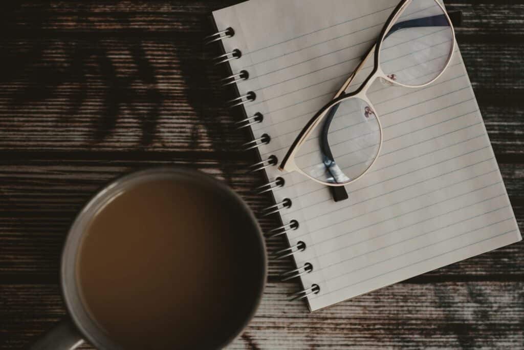 A notebook, glasses and a cup of coffee on a dark wooden table.