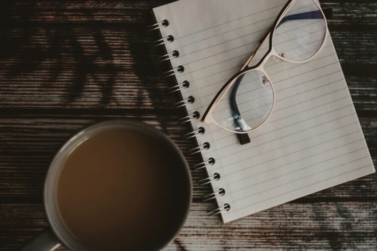 A notebook, glasses and a cup of coffee on a dark wooden table.